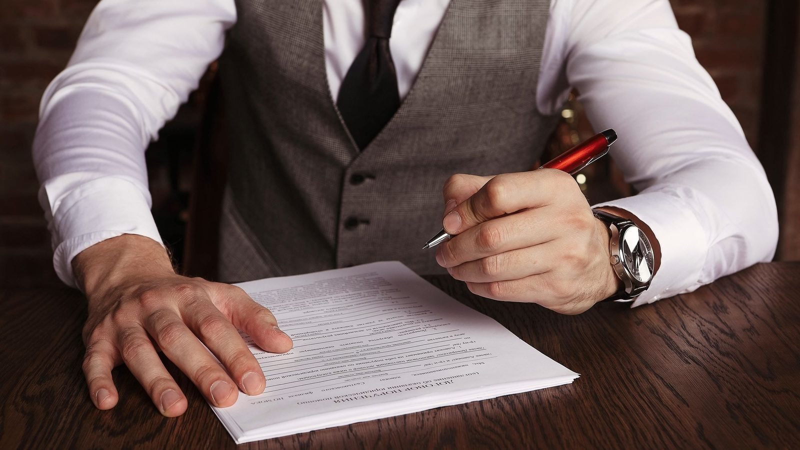 Man in a waistcoat signing a document at a dark wooden desk