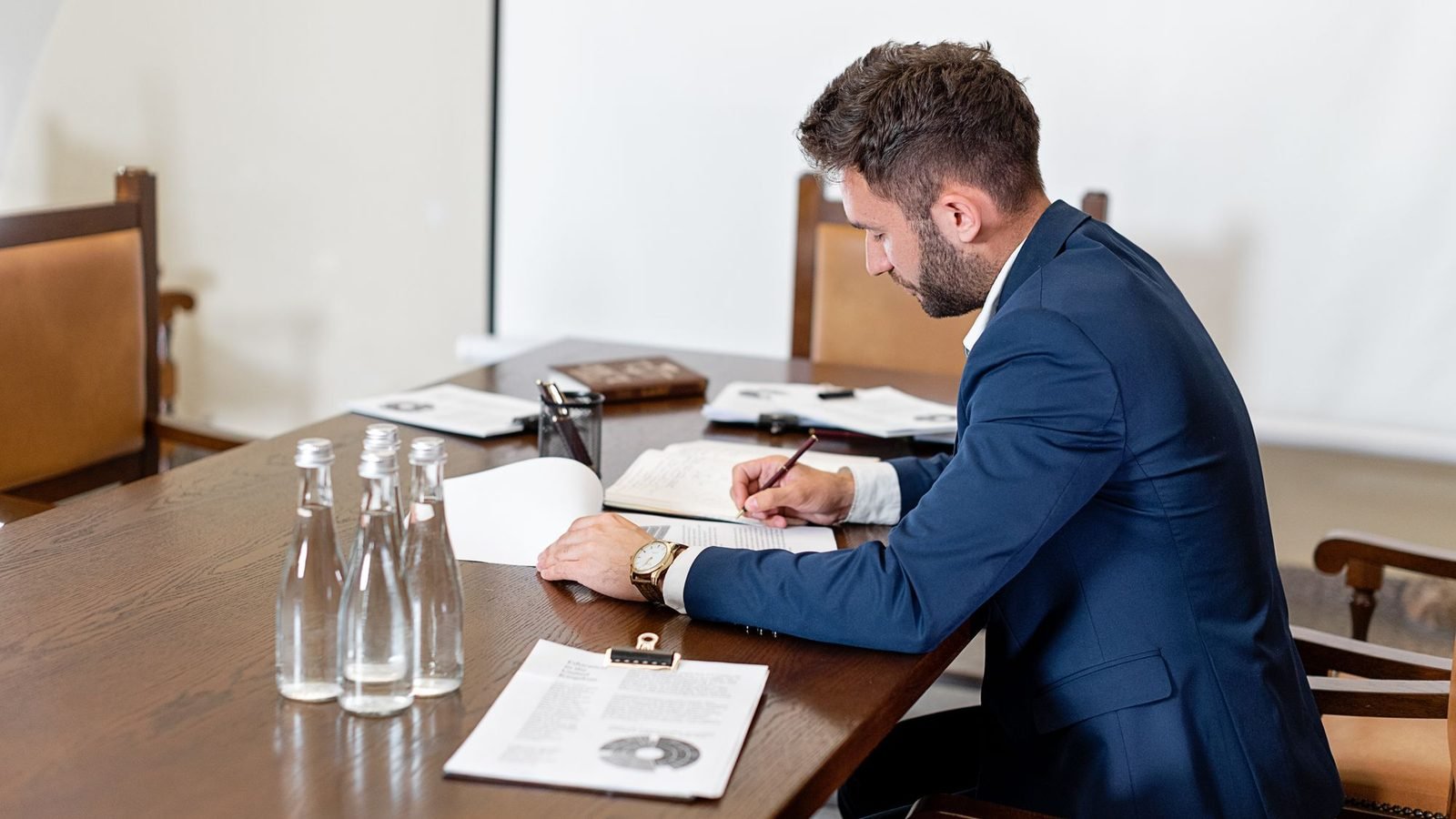 Man in a blue suit signing paperwork at a long office table