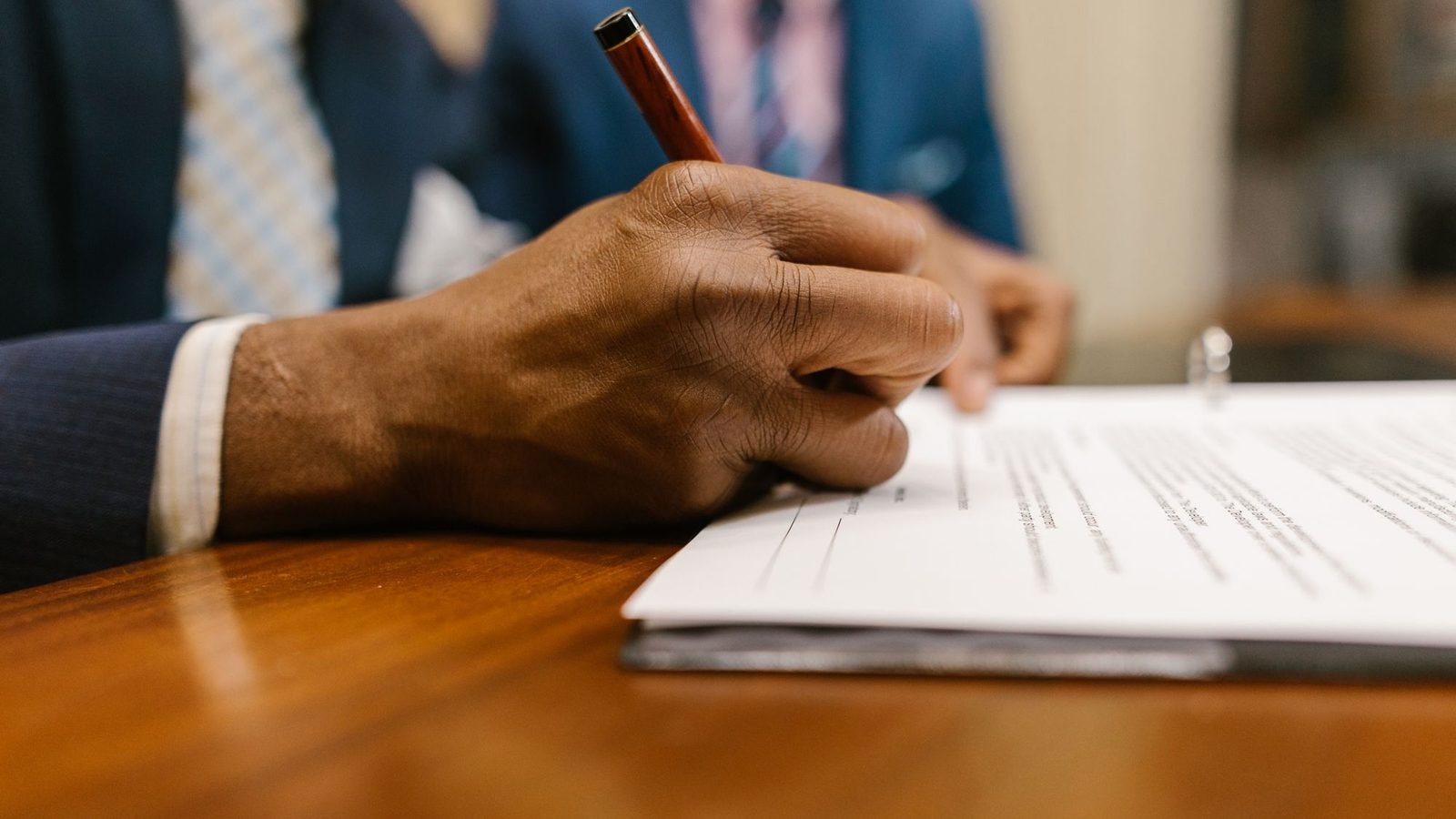 Man in a suit signing a document at a wooden desk in an office
